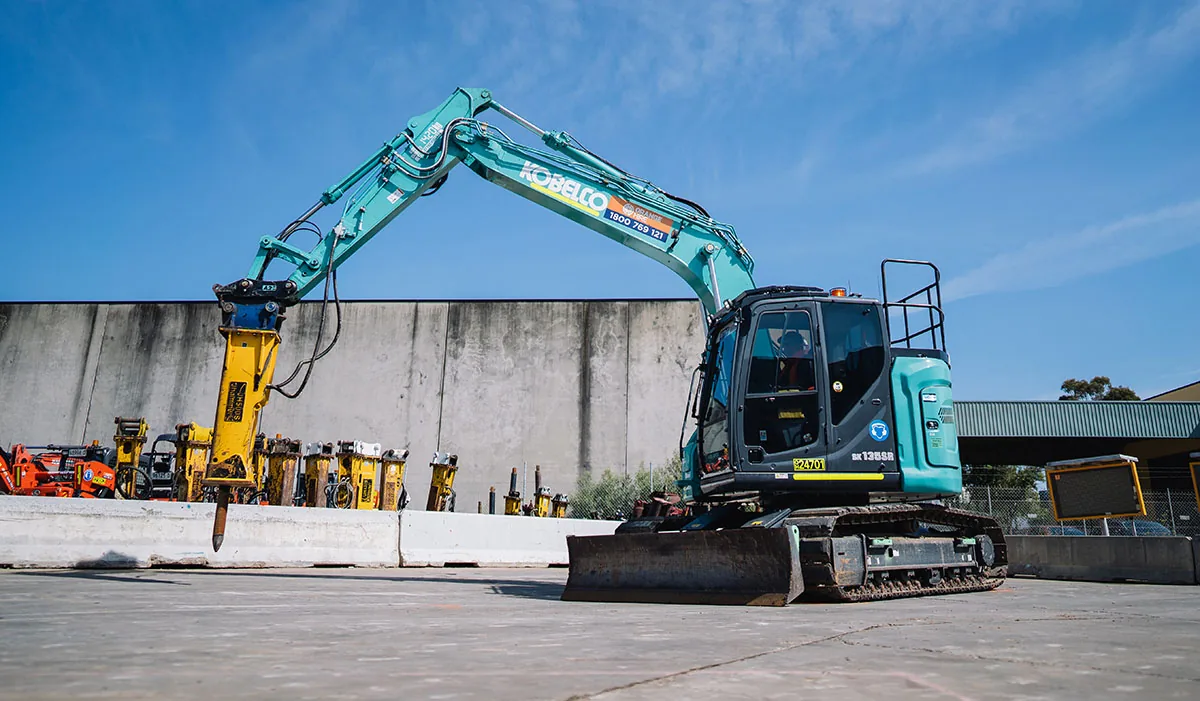 Teal blue tracked Kobelco excavator with a rock breaker attachment at a United Rentals hire yard