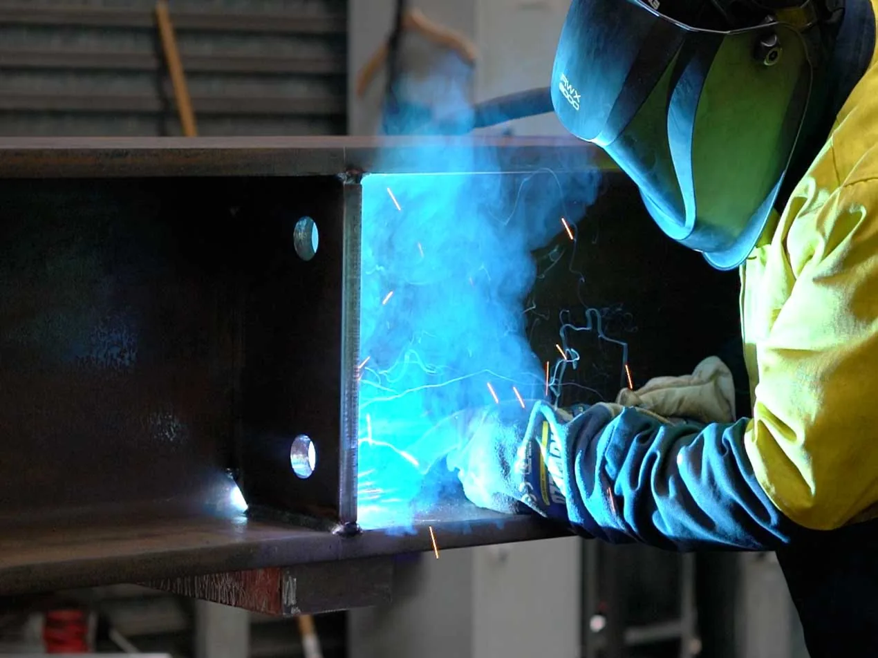 A welder in protective gear creating a bright blue arc while fabricating a large metal beam.