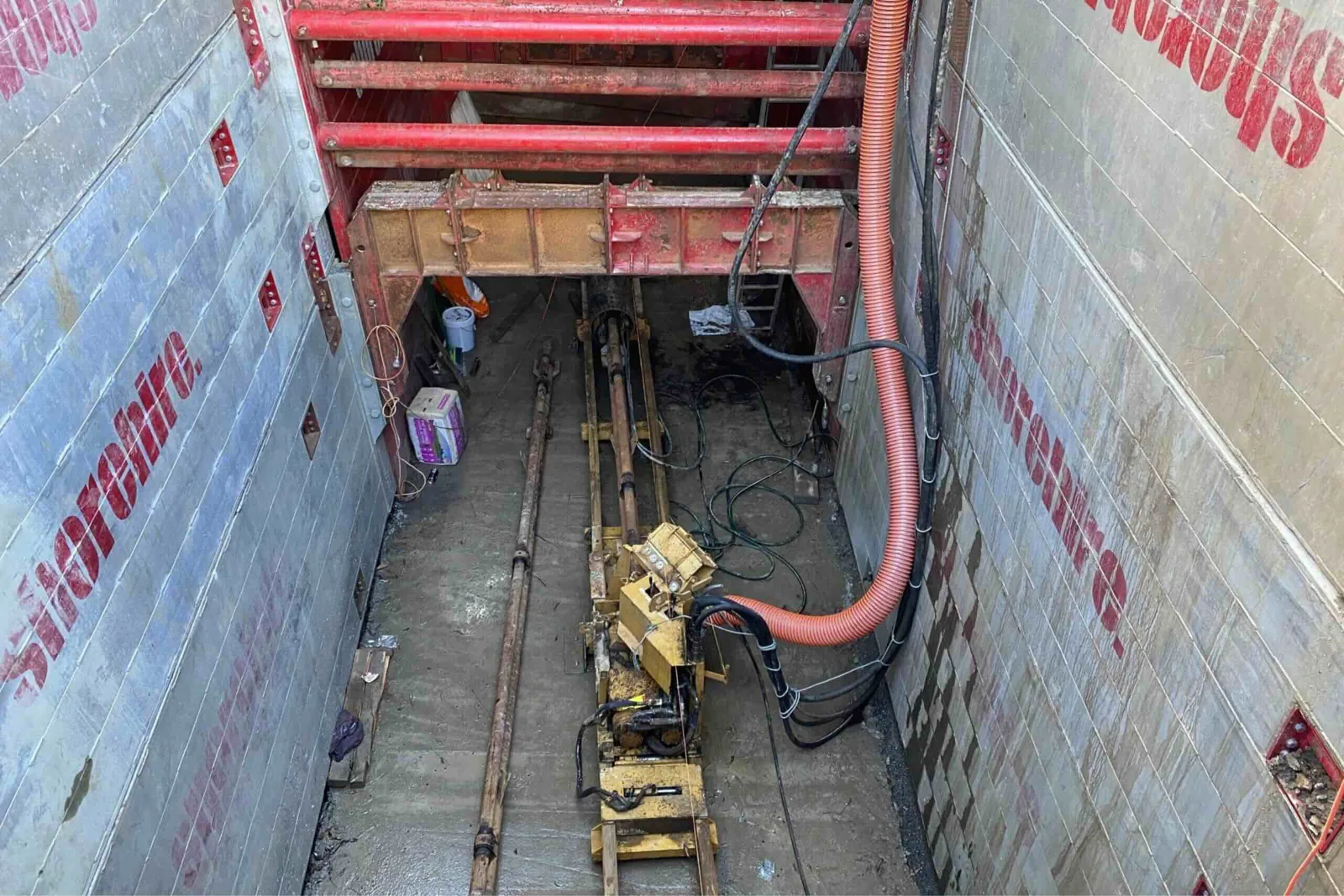 A deep trench supported by steel shoring panels contains drilling equipment and pipes arranged along the excavation floor.