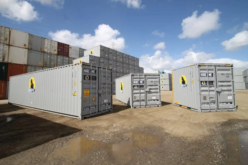 Shipping containers arranged in an outdoor yard with stacked containers in the background under a partly cloudy sky.