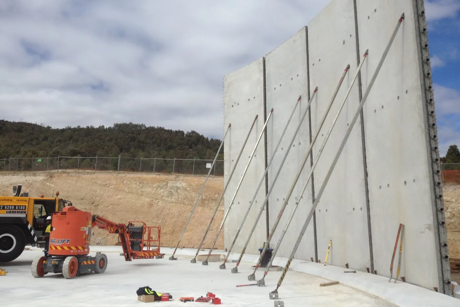 Multiple long brace supports stabilize a large precast concrete wall panel at a construction site with machinery and workers nearby.