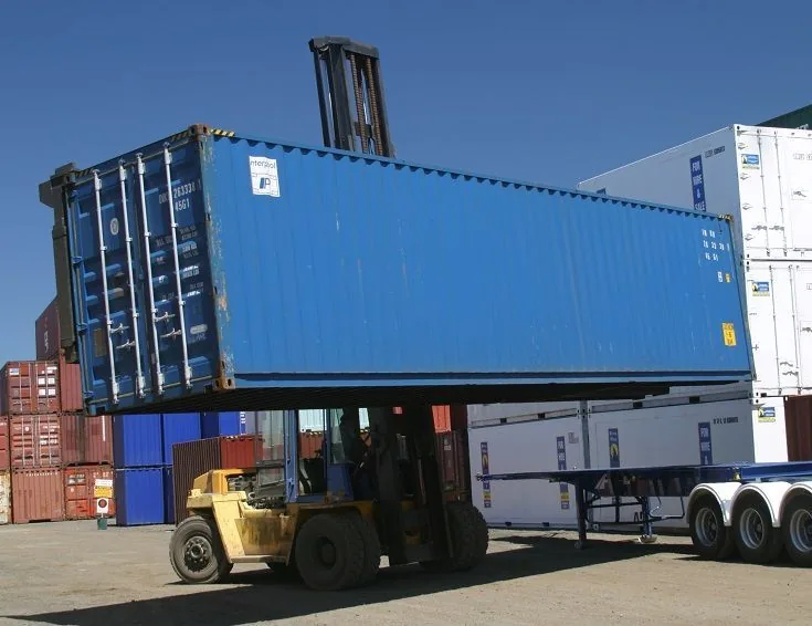 A large yellow forklift lifting a blue shipping container onto a flatbed trailer in a busy container yard.