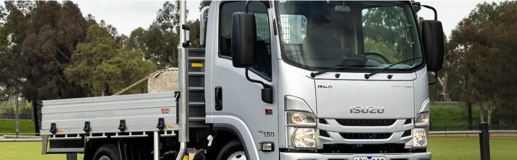 A white Isuzu flatbed truck parked outdoors, carrying a load of gravel, with trees and grass in the background.