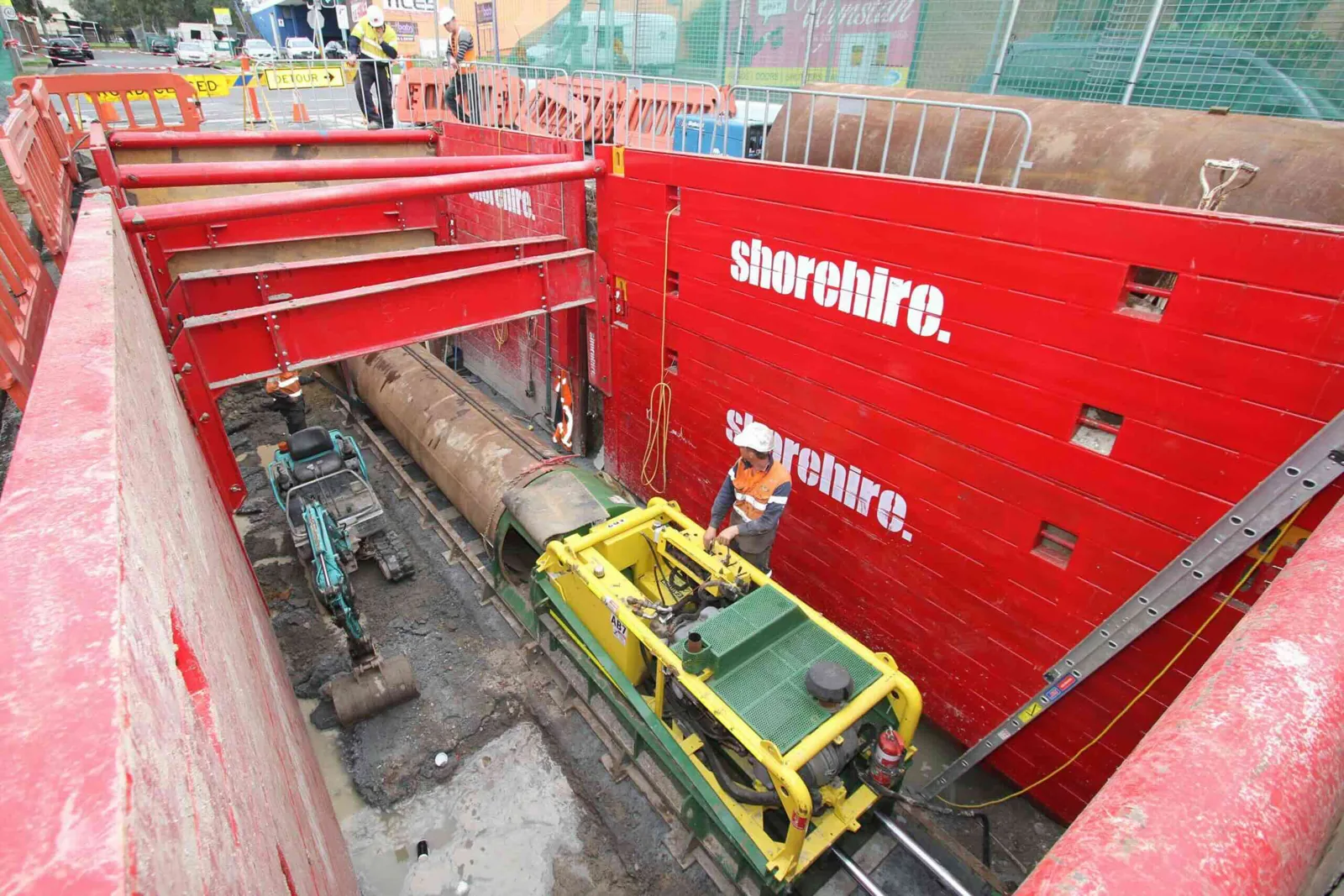 Workers operate machinery inside a large shored excavation with bright red trench boxes, surrounded by barriers and construction activity above.