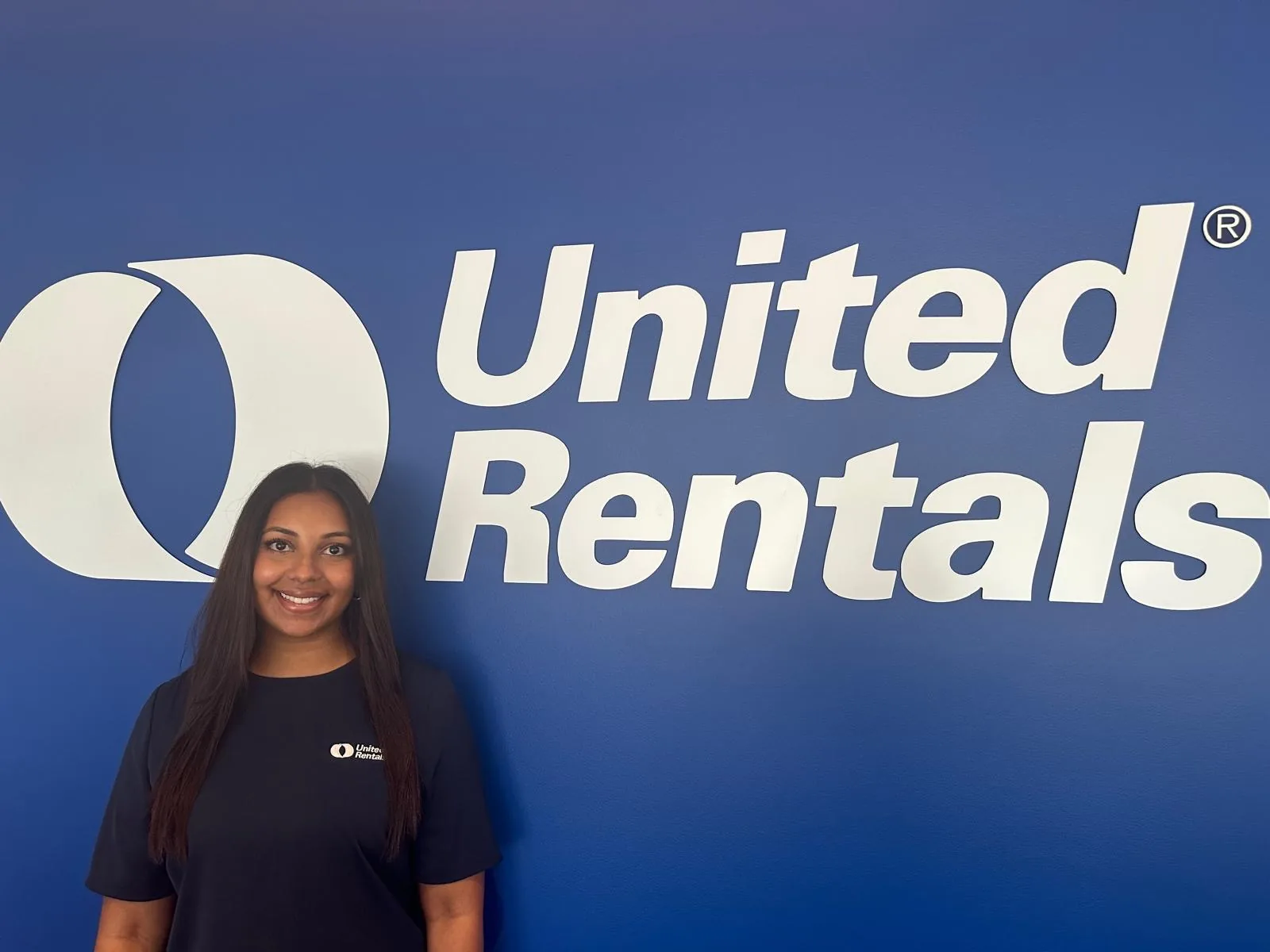 A woman standing in front of a blue wall with a big United Rentals logo on it.