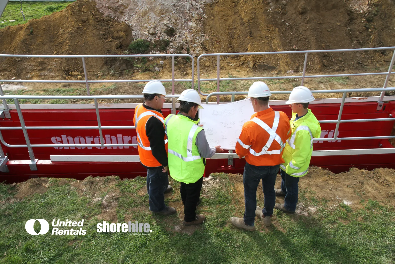 Four engineers, wearing their safety equipment, review blueprints for an excavation project while standing in front of a Steel Shoring Box equipped with an Edge Protection System, showcasing one of our Temporary Works Solutions services.