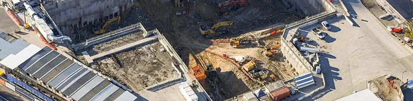 Various construction equipment working along a riverbank to build and reinforce a railway line with safety barriers.