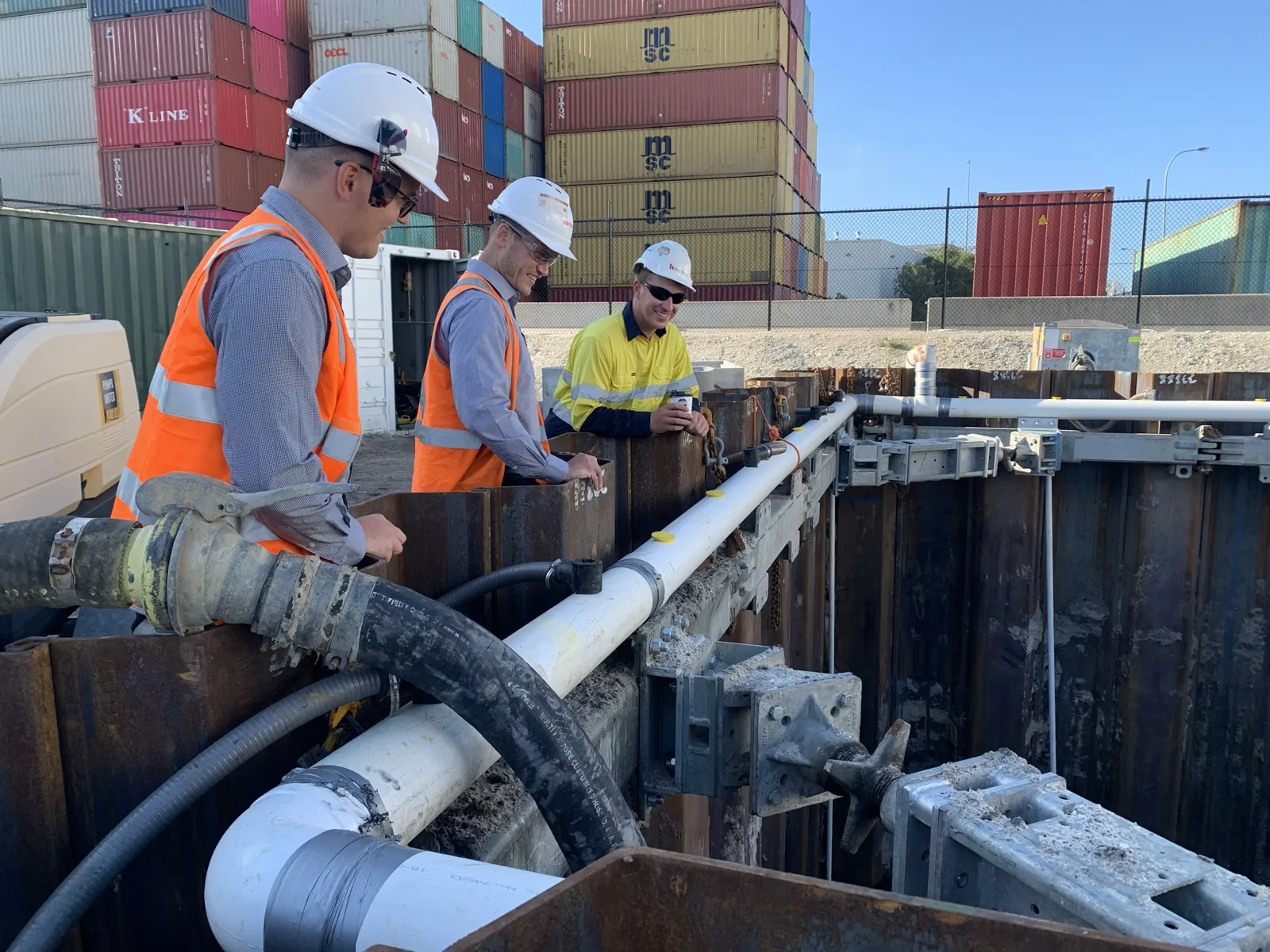 Three construction workers inspecting shoring and piping within a trench at an industrial site with shipping containers.