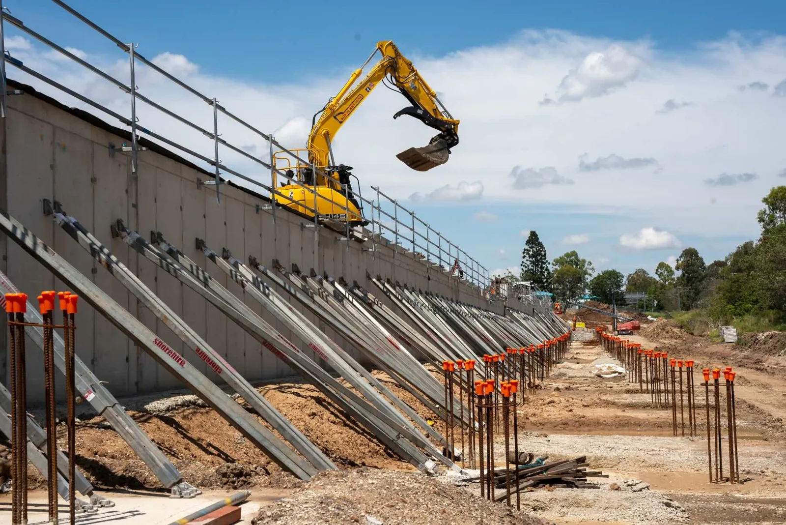 A row of angled steel brace props supports a long retaining wall while an excavator operates along the top edge of the structure.