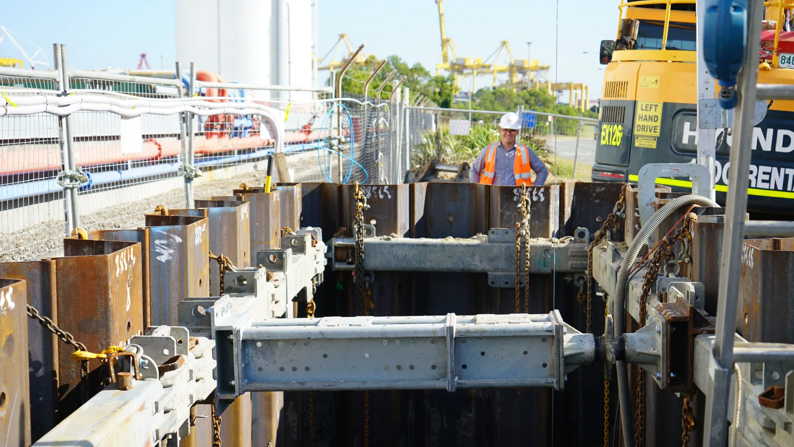 A construction worker in a hard hat and safety vest stands by a deep excavation, featuring heavy-duty metal shoring and rigging components, indicating complex installation work.