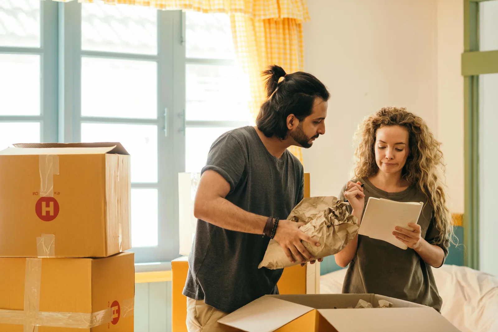 A couple packing or unpacking moving boxes, with one person holding crumpled paper and the other checking a list, suggesting a relocation process.
