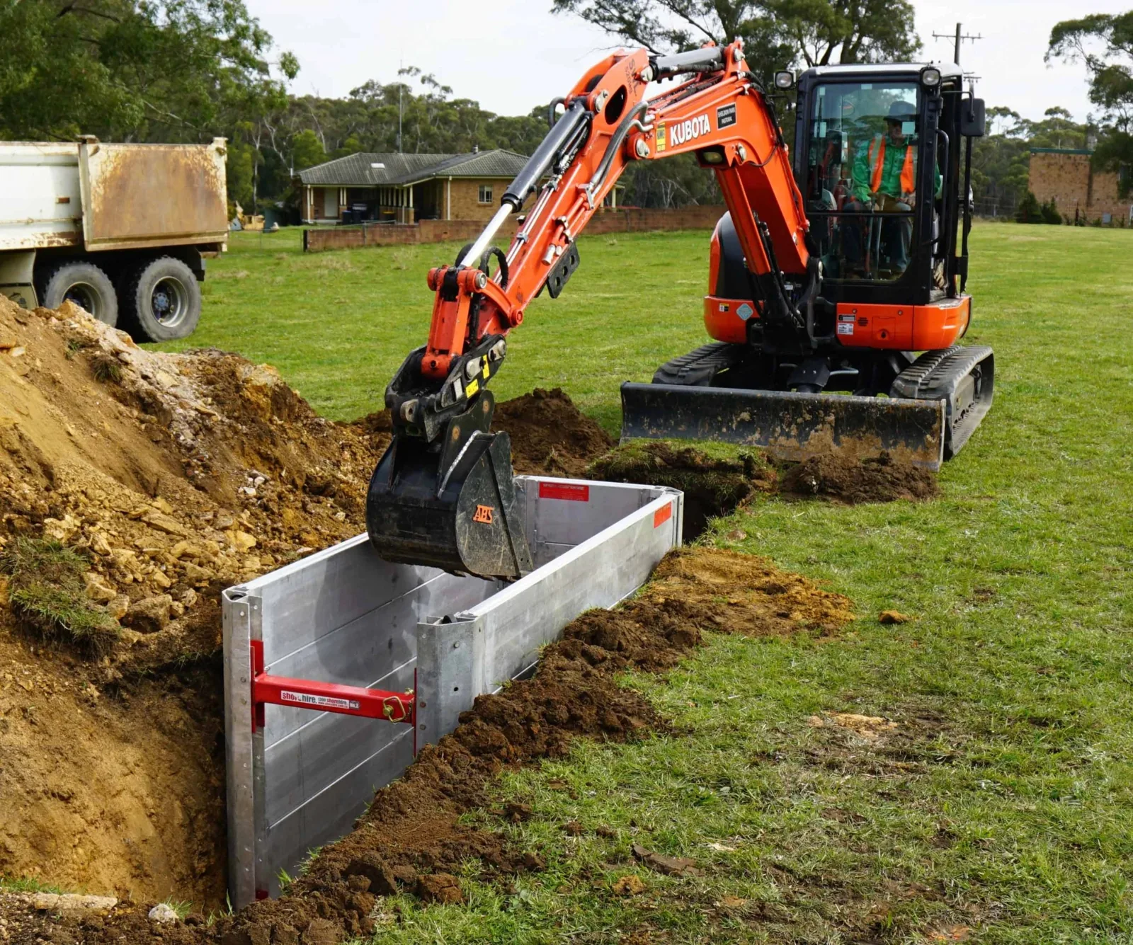 Worker operating excavation machinery near braced trench with visible hazardous material signs on a construction site.